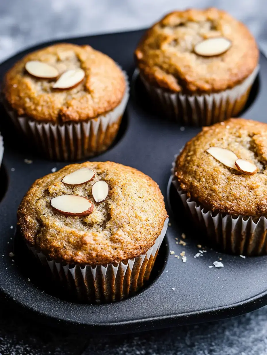 Four freshly made almond muffins sit in a dark muffin pan, garnished with sliced almonds on top.