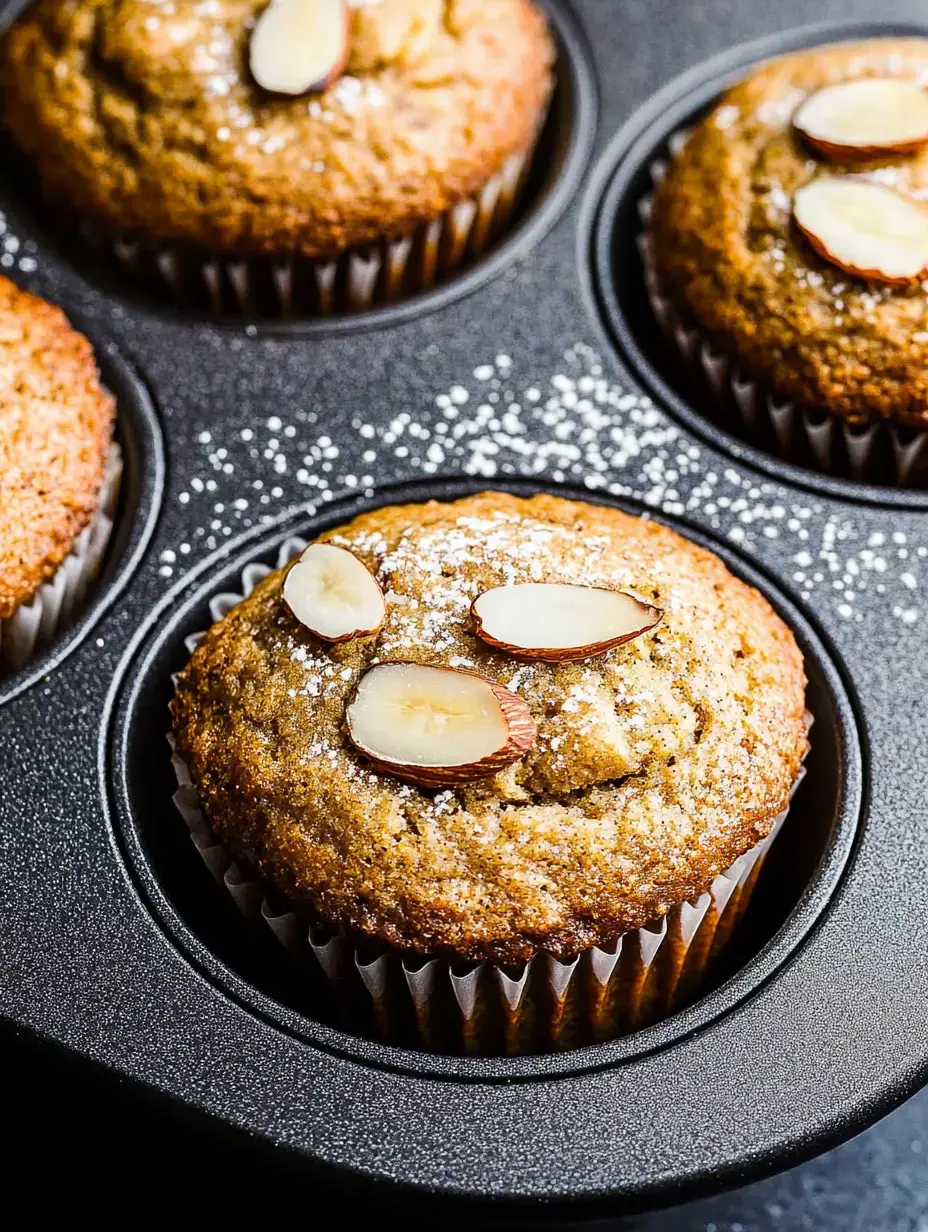 A detailed view of just-baked muffins with almond slices and sugar dusting on top, placed in a dark baking tin.